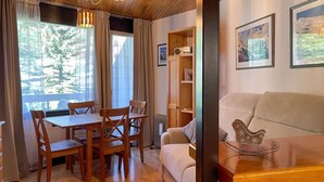 A cozy living room with a wooden ceiling, a dining area with wooden chairs and a table, a sofa, and a bookshelf.