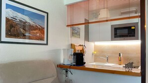 A compact hotel room with a bed, a small kitchenette with a coffee maker, a sink, and a microwave. There is a framed picture on the wall and a window with blinds.