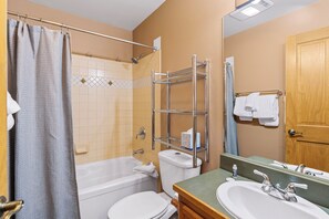 Et badeværelse med badekar, toilet og håndvask. Der er hylder over håndvasken og en bruseforhæng.