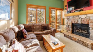 A living room with a stone fireplace, a large flat-screen TV mounted above it, and a sofa with cushions. - River Run Condo, Walking Distance to Keystone Base! Patio with Slope Views (Keystone)