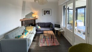 A modern living room with a grey sofa, a wooden coffee table, and a colorful rug. There's a fireplace with a lamp, a TV on a small table, and a large window with a view of the outdoors. - Renovated house with garden, wifi, BBQ, parking, 250m from the sea - Pleumeur-Bodou (Pleumeur-Bodou)