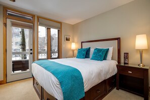 A bedroom with a large bed, two bedside lamps, a wooden nightstand, and a view through a glass door. - TR1104 | Updated Corner Unit | Winter Shuttle | 10 Hot Tubs | Close to Skiing (Steamboat Springs)