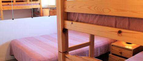 A room with two bunk beds, each with a wooden frame and a purple bedspread. There is a wooden nightstand with a lamp and a drawer. The room has a simple, functional design with a plain wall and a window.