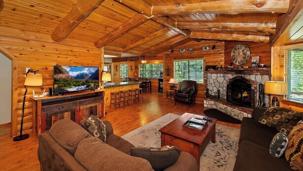 A log cabin interior with a stone fireplace, wooden ceiling beams, a television, and a bar area.