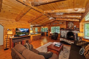 L’intérieur d’une cabane en rondins, avec une cheminée en pierre, des poutres apparentes en bois au plafond, un téléviseur et un coin bar.