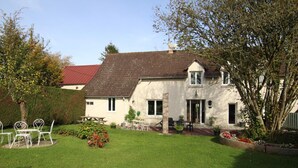 Une maison à deux étages avec une pelouse bien entretenue, un espace extérieur aménagé pour se détendre et un jardin où poussent différentes plantes et fleurs.