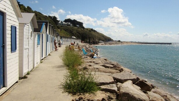Une rangée de cabanes de plage en bois peintes en blanc, avec des volets bleus, un sentier sablonneux et une côte rocheuse.