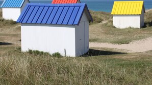 Une rangée de petites cabanes de plage à un seul étage, avec des toits en tôle ondulée peints en bleu, en rouge et en jaune, se dressant sur fond de paysage côtier.
