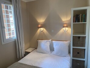 Quarto com cama, duas luminárias na parede, janela com cortinas e estante de livros.