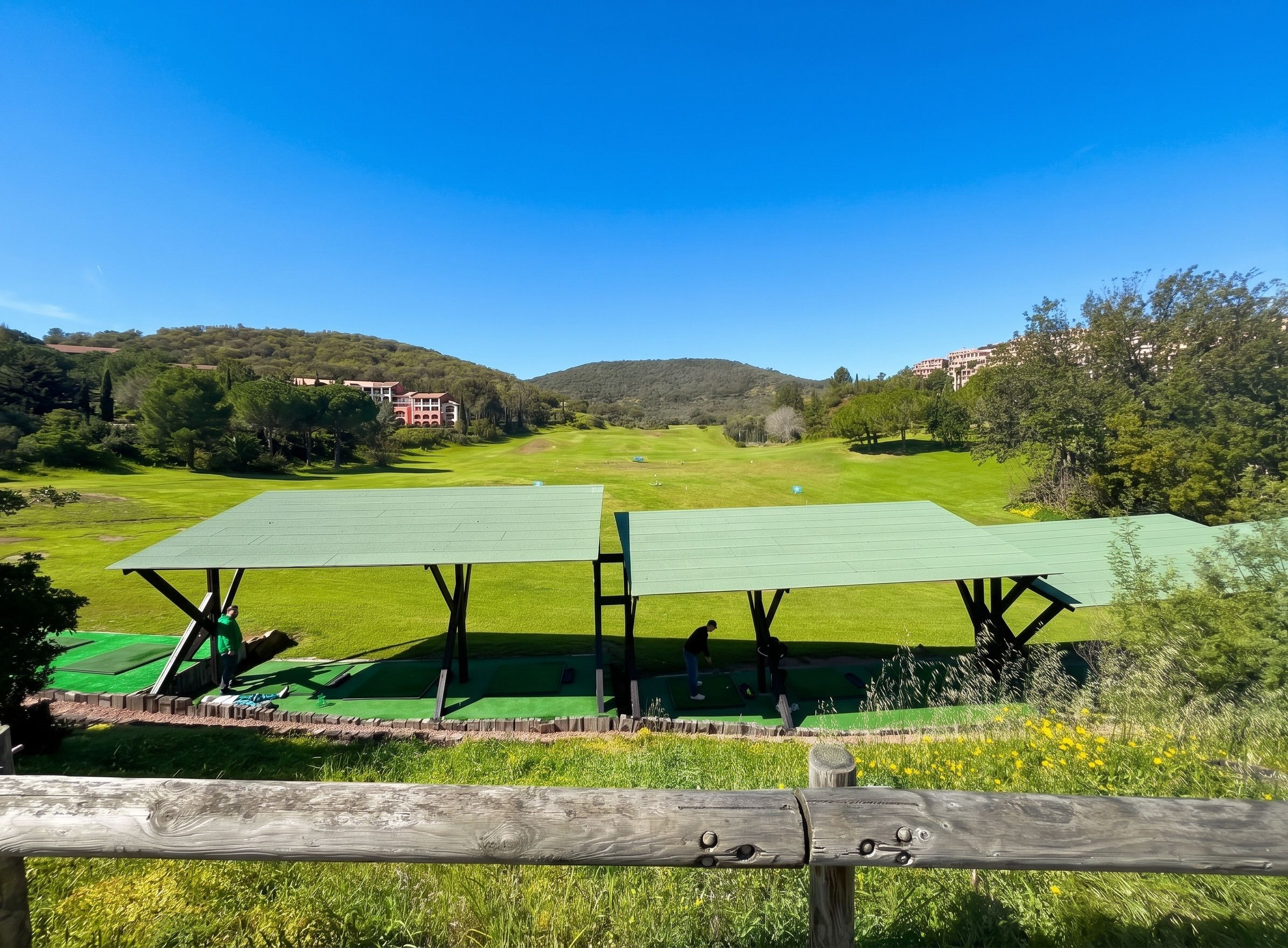 Un parcours de golf avec deux bâtiments dont les toits sont végétalisés, une clôture en bois au premier plan et un paysage vallonné en arrière-plan.