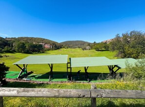 A golf course with two green-roofed structures, a wooden fence in the foreground, and a hilly landscape in the background.
