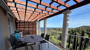 Un balcon avec une table et des chaises, un barbecue et une vue sur la mer et la verdure.