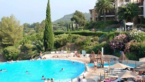 Un espace piscine où des personnes nagent et prennent des bains de soleil, entouré de verdure et bordé d’un bâtiment doté de balcons.