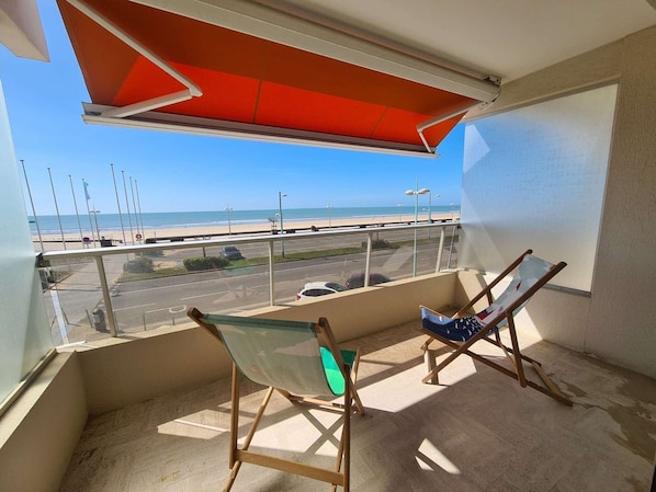Un balcon avec deux fauteuils de jardin en bois, un auvent rouge et une vue sur la plage et le parking.