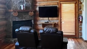 A log cabin interior with a stone fireplace, a flat-screen TV mounted on a wooden wall, and two leather recliner chairs. - River Alpine Loft managed by Cabin Butler (Greer)