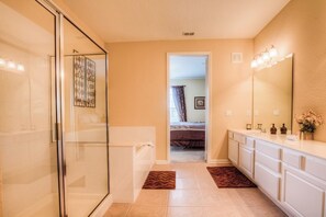 A bathroom with a glass shower enclosure, a bathtub, a vanity with a mirror, and a door leading to a bedroom.