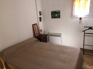 A bedroom with a bed, a radiator, a small table with a lamp, and a window with curtains.