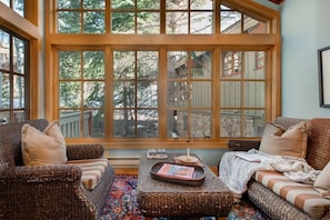 Une pièce chaleureuse, meublée de meubles en osier, avec une grande fenêtre donnant sur les arbres et un tapis à motifs.