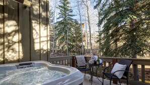 A hot tub on a wooden deck with chairs and a table, surrounded by trees.