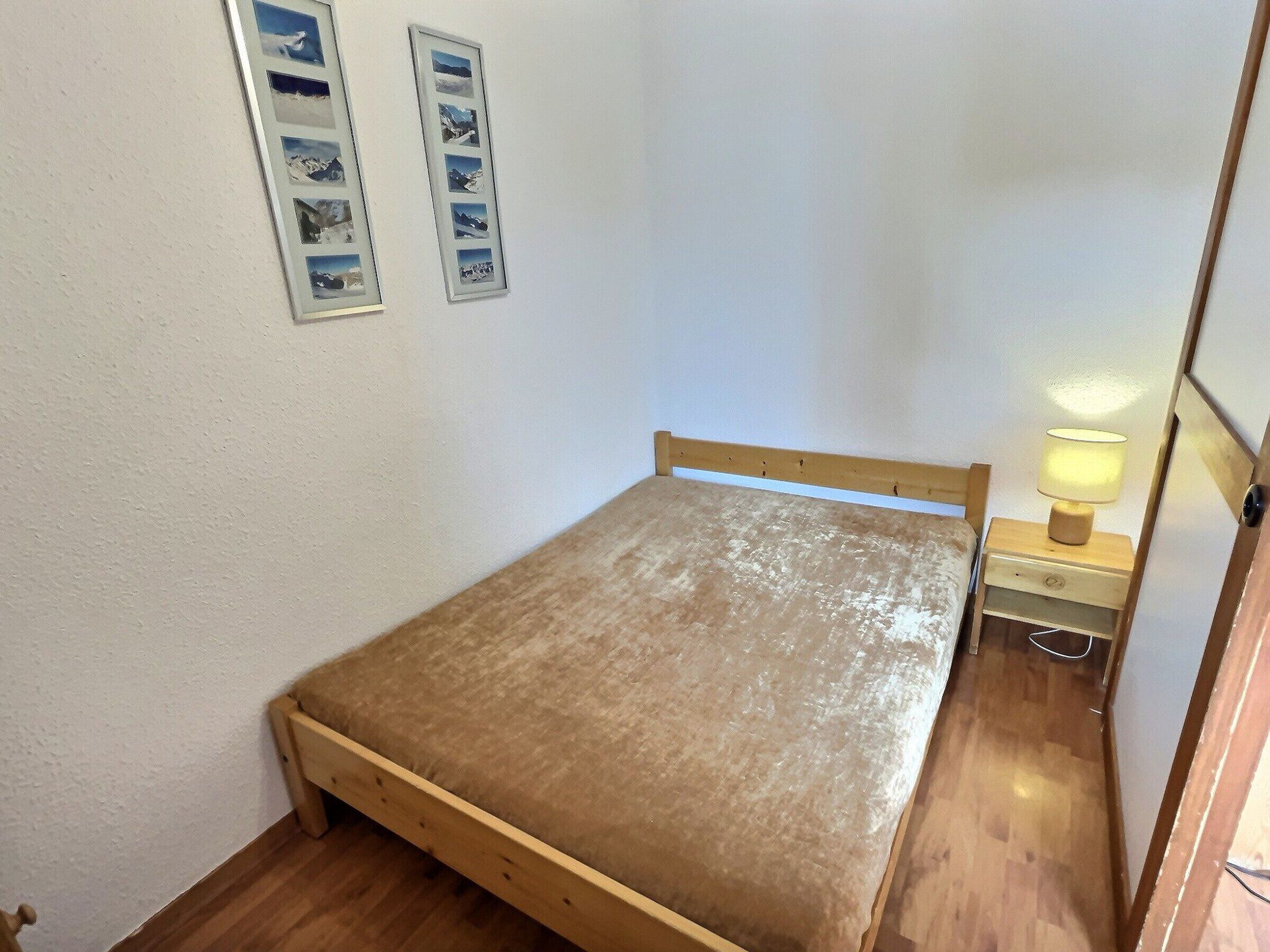 Et lille værelse med en enkelt seng, sengegavl i træ, et natbord med lampe og to billeder i rammer på væggen.
