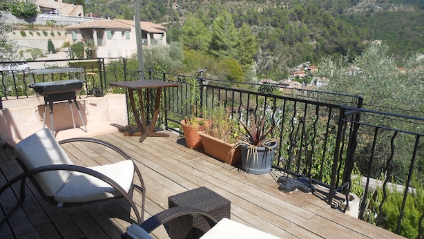 A wooden deck with two wicker chairs and a table, surrounded by potted plants and a metal railing. - RENTAL GITE BELOW VILLA (L'escarène)