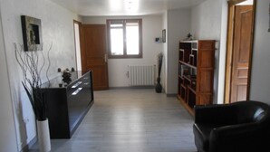 A spacious living area with a wooden floor, a black sofa, a bookshelf, and a dining area with a black table and chairs. - RENTAL GITE BELOW VILLA (L'escarène)