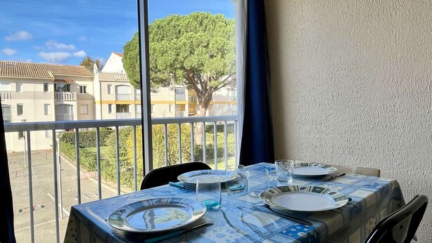 A balcony with a table set for dining, overlooking a residential area with trees and buildings. - Cozy Studio Near Beach, Shops, Private Garage, Pet-Friendly, Sleeps 4 (Argelès-sur-Mer)