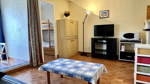 A compact living space with a bunk bed, a television, a microwave, and a small table with a patterned tablecloth. - Cozy Studio Near Beach, Shops, Private Garage, Pet-Friendly, Sleeps 4 (Argelès-sur-Mer)