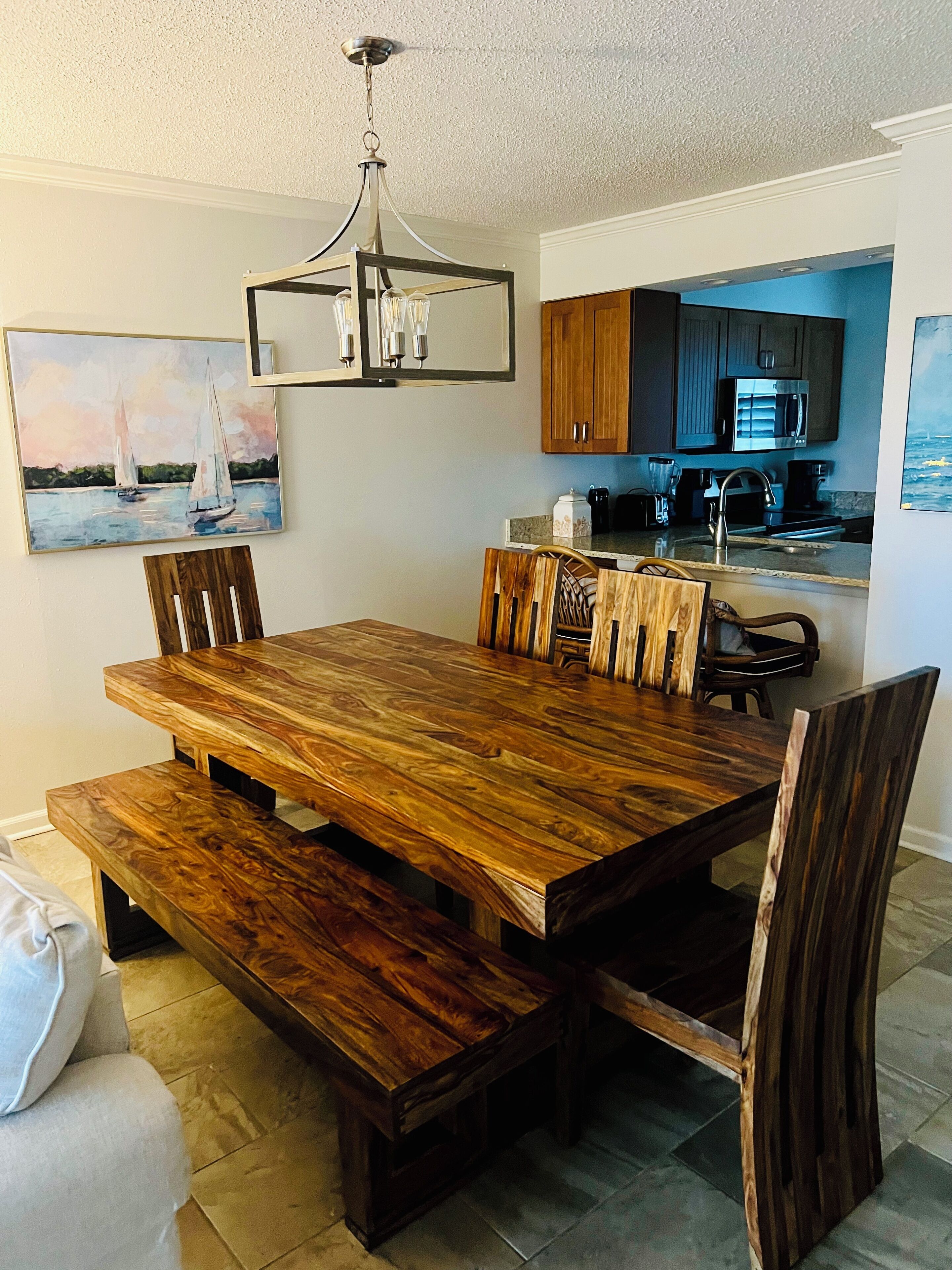 A dining area with a wooden table and chairs, a chandelier, and a kitchen in the background.