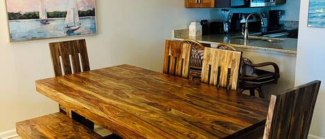 A dining area with a wooden table and chairs, a chandelier, and a kitchen in the background.