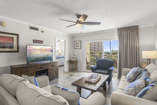 A spacious living room with a flat-screen TV, comfortable seating, a ceiling fan, and a view through large sliding doors to an outdoor balcony.
