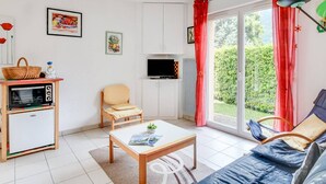 A living room with a sofa, a small table, a TV, a chair, a basket, and a window with curtains. - Calm T3 for 6, terrace, garden, near amenities (Esquièze-Sère)