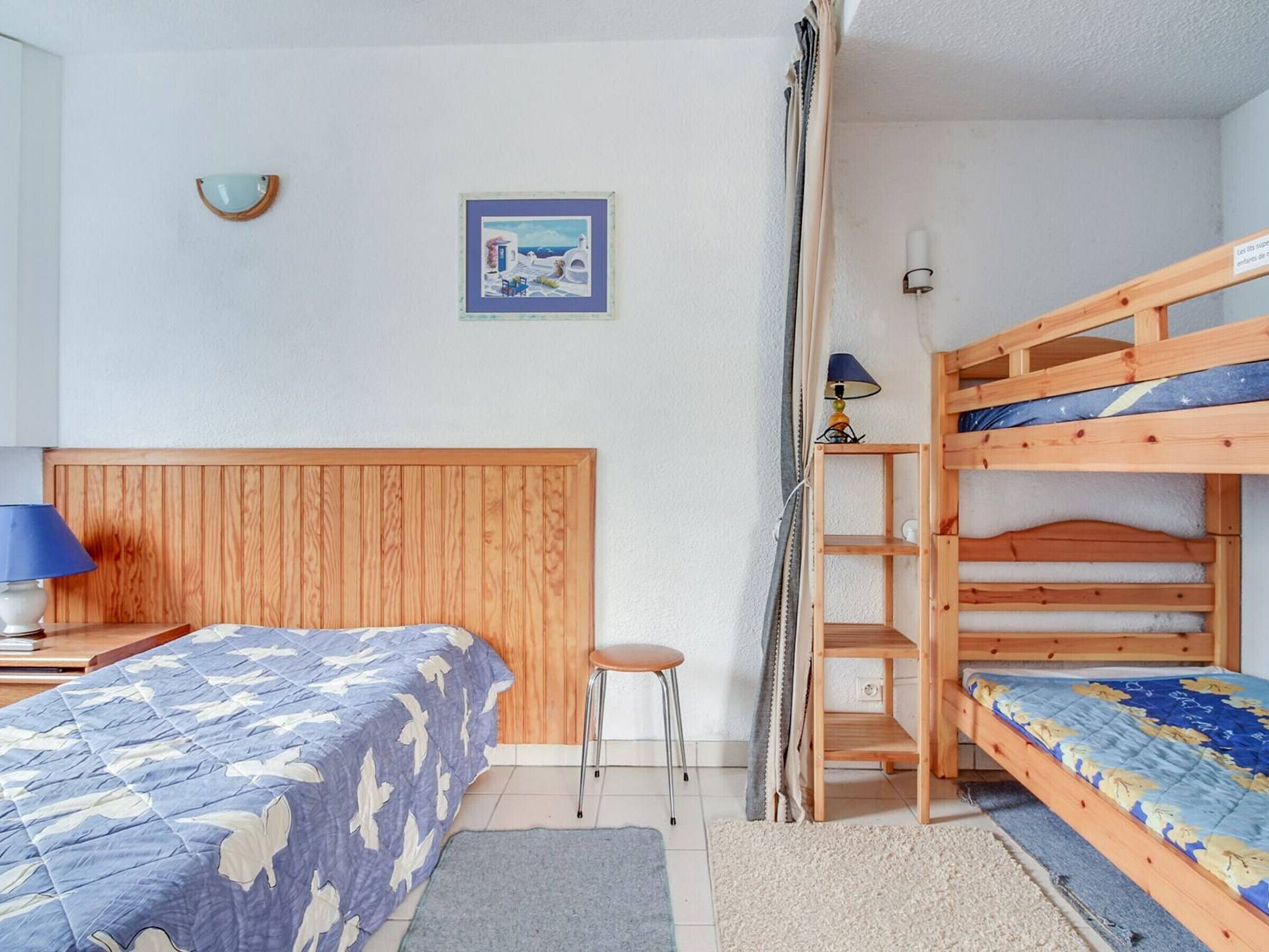 A bedroom with a bunk bed, a single bed, a small stool, a lamp, and a framed picture on the wall.