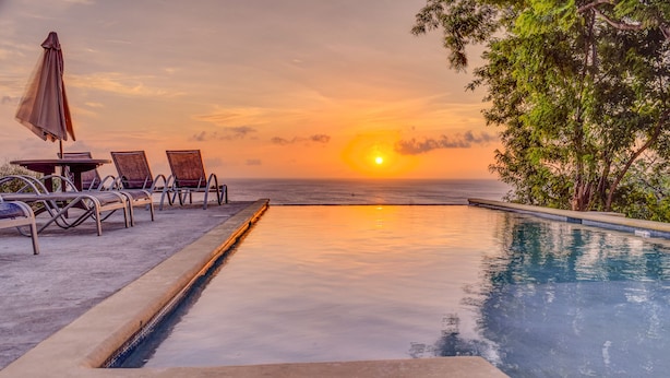 Ein Poolbereich mit Liegestühlen und einem Sonnenschirm, mit Blick auf den Ozean bei Sonnenuntergang.