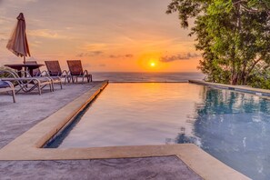 A poolside area with lounge chairs and an umbrella, overlooking the ocean at sunset. - New Luxury 1 Bedroom SeaScape Condo in West Roatan w/ Pool & Ocean Views (West Bay)