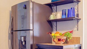 Eine kompakte Küche mit einem Edelstahl-Kühlschrank, einem Einbauofen und einem Regal darüber, das blaue Tassen und einen Obstkorb beherbergt.
