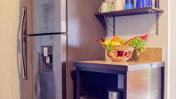Una cocina compacta con un refrigerador de acero inoxidable, un horno empotrado y una repisa con tazas azules y una cesta de fruta.