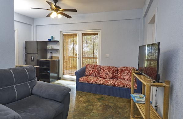 A living room with a sofa, a television, a ceiling fan, and a sliding glass door leading to a balcony. - New Luxury 1 Bedroom SeaScape Condo in West Roatan w/ Pool & Ocean Views (West Bay)