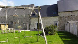 Un jardin avec une balançoire en bois, un bâtiment en pierre et une zone délimitée par une clôture.