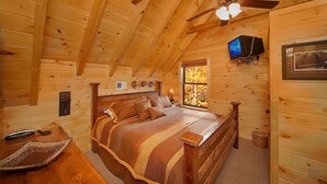 Un dormitorio rústico tipo cabaña de madera, con cama de madera, ventilador de techo, televisor montado en la pared y ventana con follaje otoñal visible en el exterior.