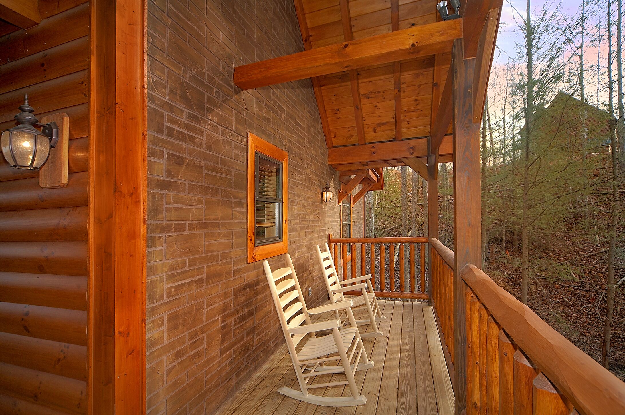 Una terraza de madera con sillas mecedoras, un muro de ladrillo y una barandilla de madera que da a un bosque.