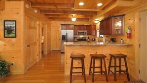 Interior de una cabaña de madera con una zona de cocina que incluye un refrigerador, microondas y estufa. Hay tres taburetes de madera en la encimera de la cocina.