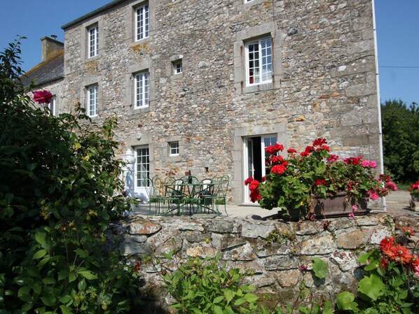 A stone building with multiple windows and a door, surrounded by greenery and flowers.