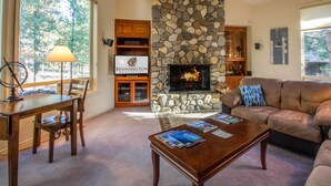 A cozy living room with a stone fireplace, a wooden coffee table, a leather sofa, and a dining area with a lamp and chairs. - CYPR10 - Huge Home, 2 Suites, Dog Friendly, Hot Tub (Sunriver)