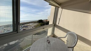 A balcony with a table and chairs, overlooking a parking area and a beach.