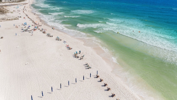 Une vue aérienne d’une plage avec des rangées de transats et de parasols, un ciel bleu dégagé et l’océan.