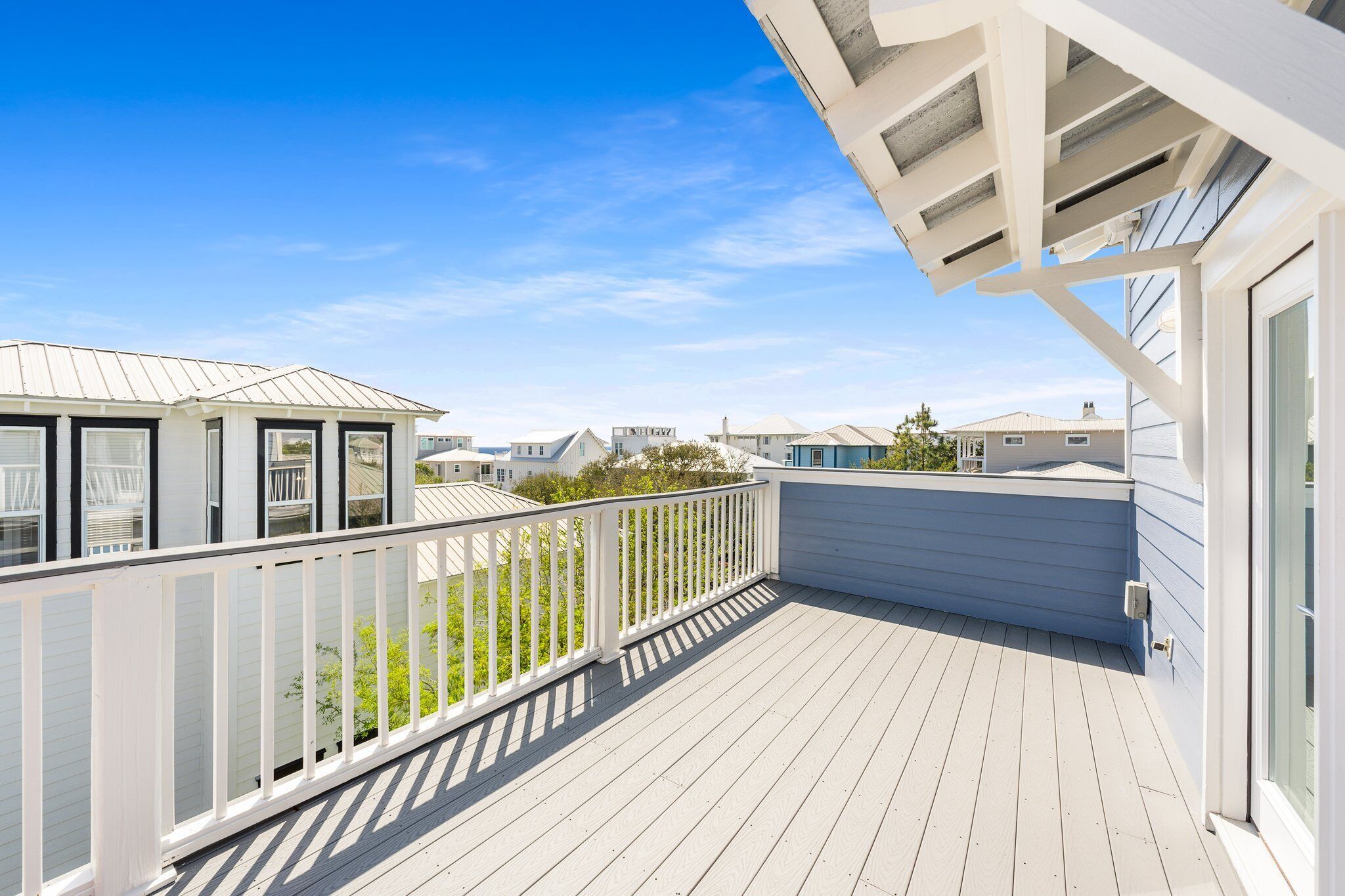 Une terrasse en bois avec une balustrade blanche, donnant sur un quartier résidentiel avec des maisons et un ciel bleu dégagé.