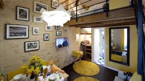 Una habitación acogedora con cama elevada, un rincón de comedor con sillas amarillas, un televisor y una pared decorada con fotografías en blanco y negro.