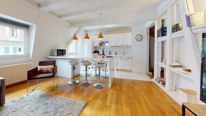 A modern kitchen with white cabinets, a microwave, and orange pendant lights. There is a dining area with bar stools and a small rug.