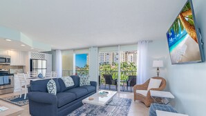 Una sala moderna con sofá, mesa de centro y sillones, un área de cocina y un balcón con vistas a palmeras y edificios.